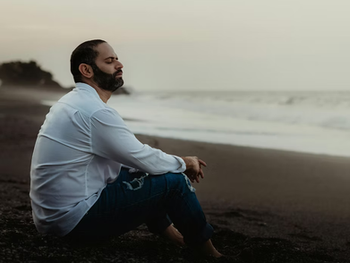 A man sitting quietly on a beach with his eyes closed, facing the ocean in a peaceful moment of reflection and contemplation.