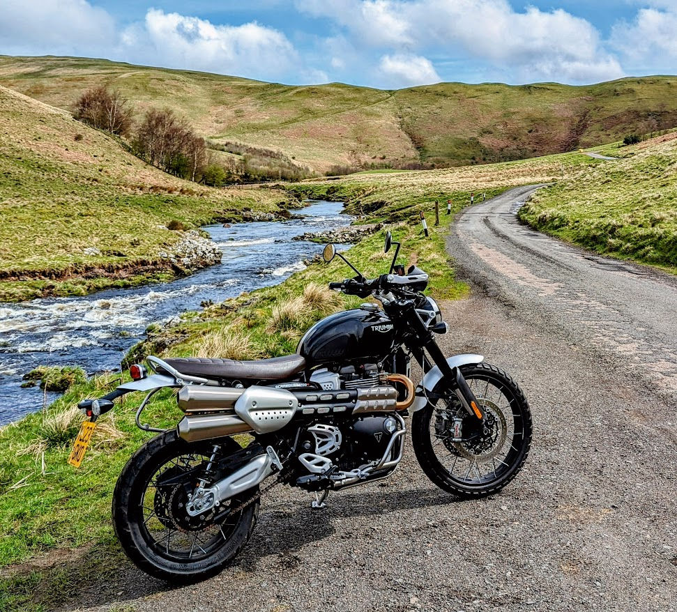 Black Triumph motorcycle parked beside a winding rural road and stream in green hills under a cloudy blue sky.