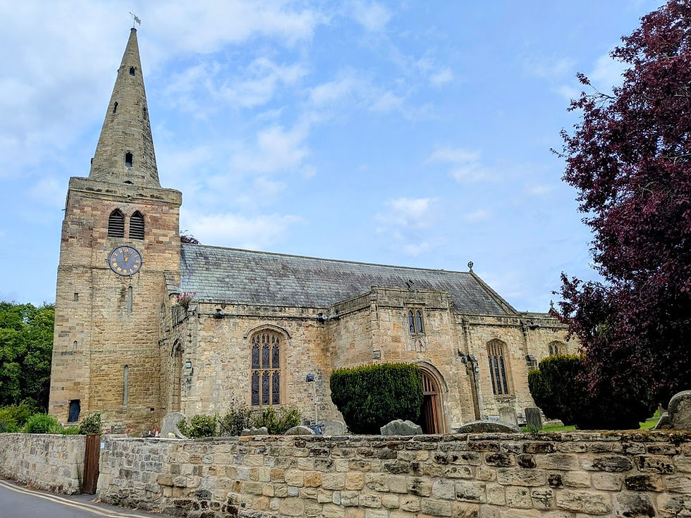 Stone church with tall spire and clock behind a low stone wall, set against a blue sky and trees.