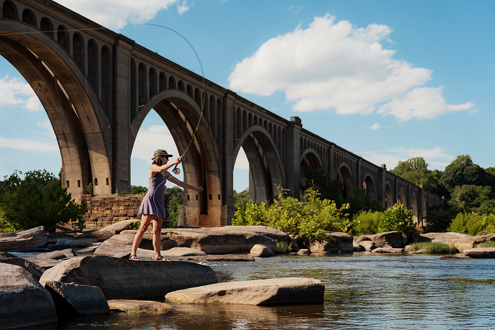 A woman casting a fly rod while standing on a rock with a bridge in the background.