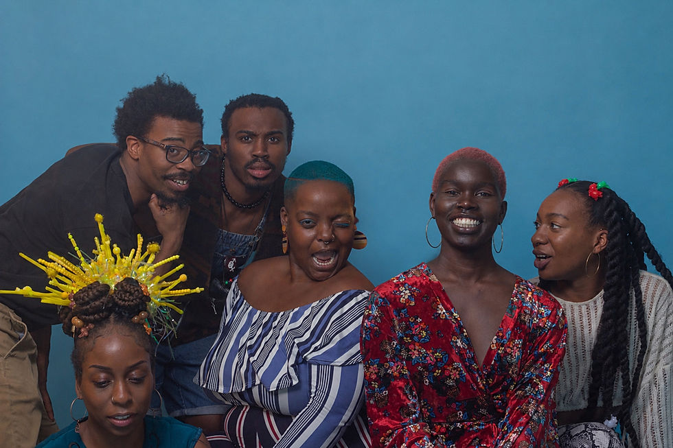 Six Black people crowd together playfully against a bright blue studio backdrop during an early Afro ABCs test shoot. Their hair includes a towering yellow sculptural arrangement built around braided buns, a teal close crop, a coral-pink crop, long twists decorated with red-and-green beads and several short natural styles.