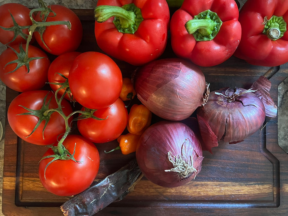  Overhead photograph of ripe red tomatoes, red bell peppers, small hot peppers, and red onions gathered on a wooden cutting board before being roasted and blended into pepper mix.
