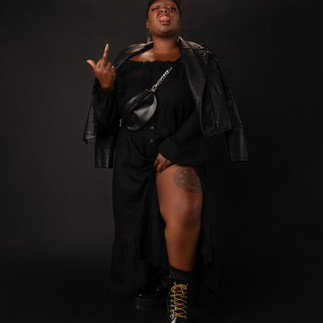 Portrait of Kiing Curry against a black studio backdrop wearing a flowing black dress, leather jacket, black cap, crossbody bag, and black platform boots with gold laces. They lift one side of the dress while “flipping a bird” to reveal a thigh tattoo while making a playful expression toward the camera during the Eloquii Black Creators Project.