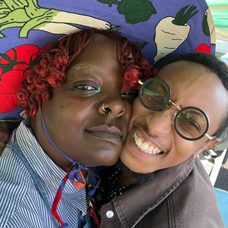 Close-up selfie of two smiling people at a Juneteenth event. One wears a vegetable-print hat and red curls while the other wears round black glasses.