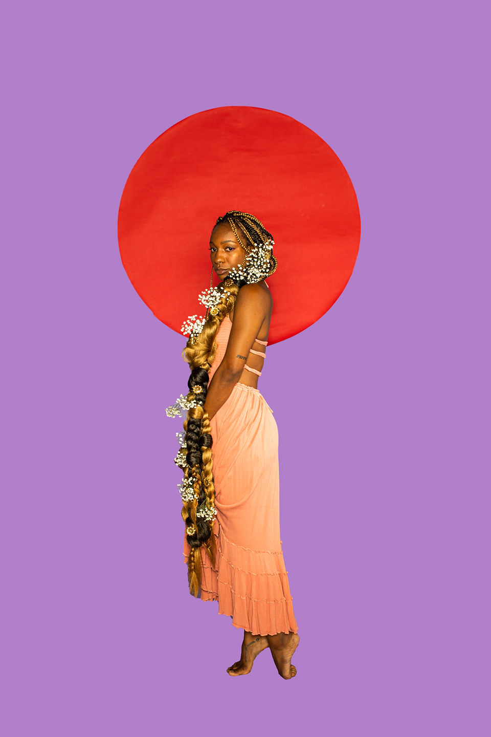 Concept portrait of a Black model portraying Rapunzel, standing against a graphic studio backdrop with an oversized red sun while an extraordinarily long braided hairstyle wrapped in flowers extends far beyond the frame. Part of the unfinished Black Fairytales series inspired by Fred Crump Jr.’s Black fairy tale books.