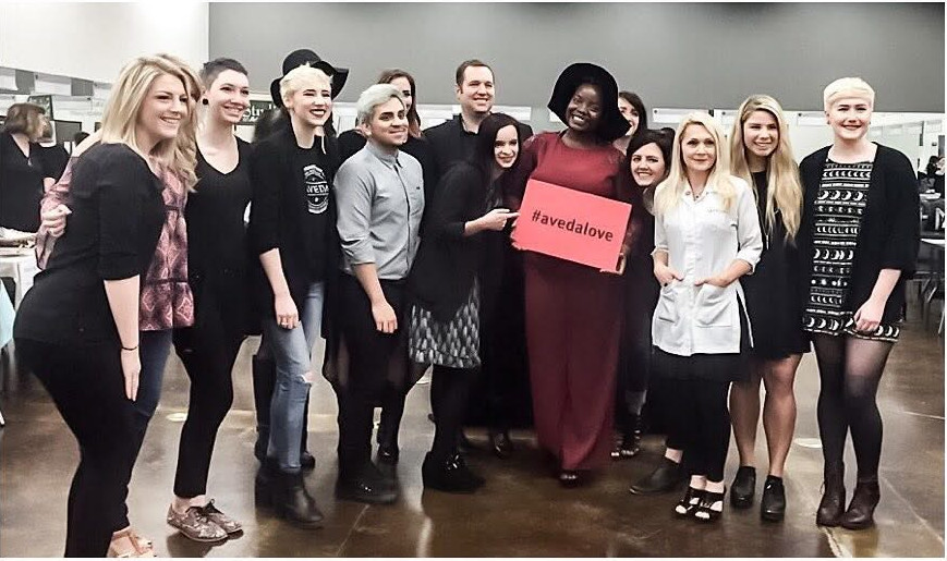 Kiing—the only Black body in the group—stands at the center of a sea of white cosmetology students and instructors. Wearing red and a wide-brimmed black hat, they hold a pink sign reading “#avedalove” while the group crowds around them inside the school.