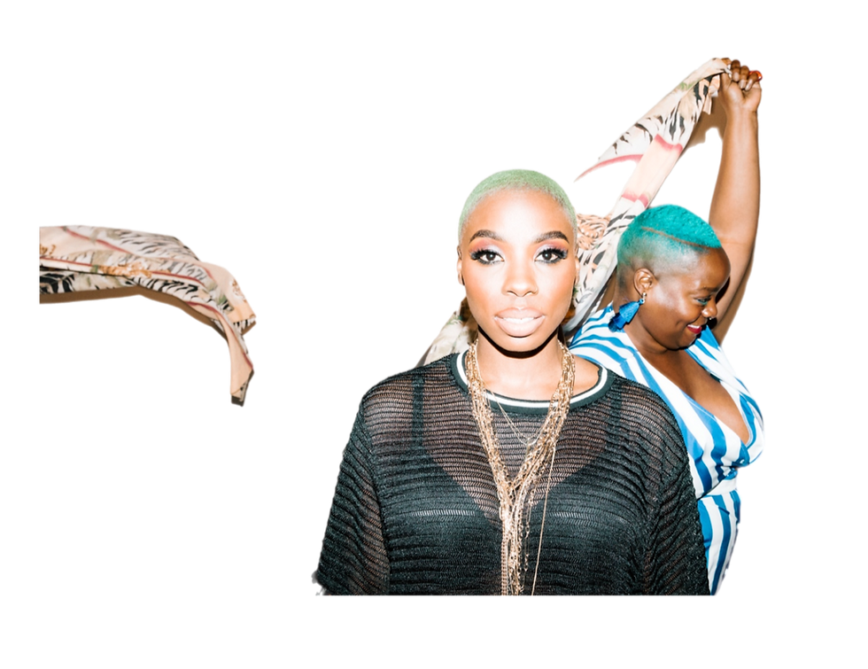 Behind-the-scenes photograph from Jessy Wilson’s first album “Phase” photoshoot. Jessy Wilson stands in the foreground with closely cropped green hair, layered necklaces, and a sheer black top while Kiing Curry smiles in the background with raised arms during a candid moment between setups.