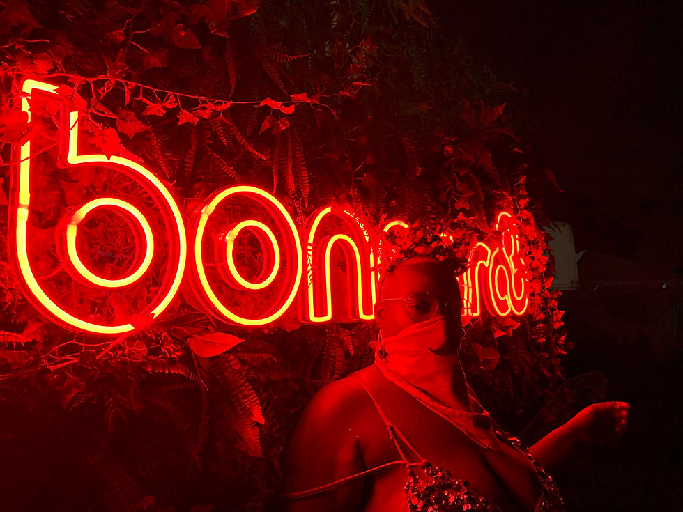 person standing with side to camera in front of bright red neon “bonnaroo” sign surrounded by foliage, body partially illuminated by red light