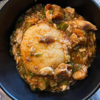 Black bowl filled with okra soup surrounding a smooth rice ball. Chopped okra, seafood, and rich orange broth surround the center, with visible strands of draw connecting ingredients throughout the dish.