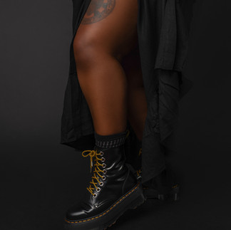 Close-up portrait of Kiing Curry’s black platform boots with gold laces, exposed thigh tattoo, and flowing black garment against a black studio backdrop during the Eloquii Black Creators Project.