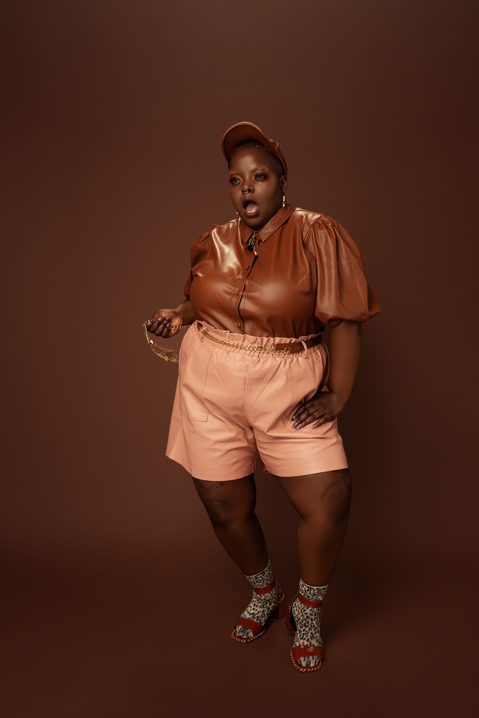 Portrait of Kiing Curry standing against a warm brown backdrop wearing a brown faux leather blouse, blush shorts, a matching cap, patterned socks, and heeled sandals while holding a chain accessory during the Eloquii Black Creators Project.