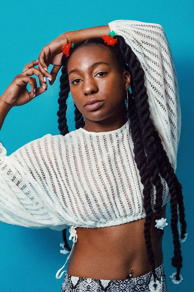 A Black model poses against a bright blue backdrop wearing a white open-knit crop top. Their hair is divided into two long bundles of thick twists, decorated at the roots with red-and-green clips and along the lengths with translucent white flowers.