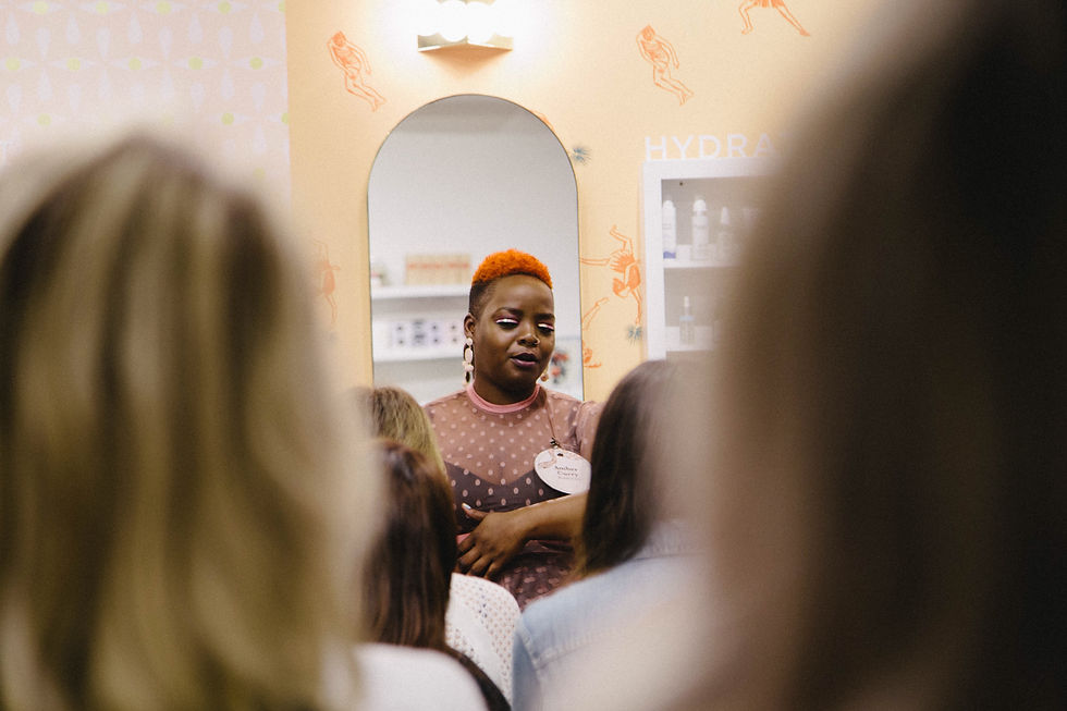 Kiing, with closely cropped orange hair and white graphic eye makeup, speaks to a gathered group inside Lemon Laine beauty space. They wear a sheer pink polka-dot top and a name badge. The blurred backs of attendees frame Kiing at the center, with an arched mirror and shelves of products behind them.
