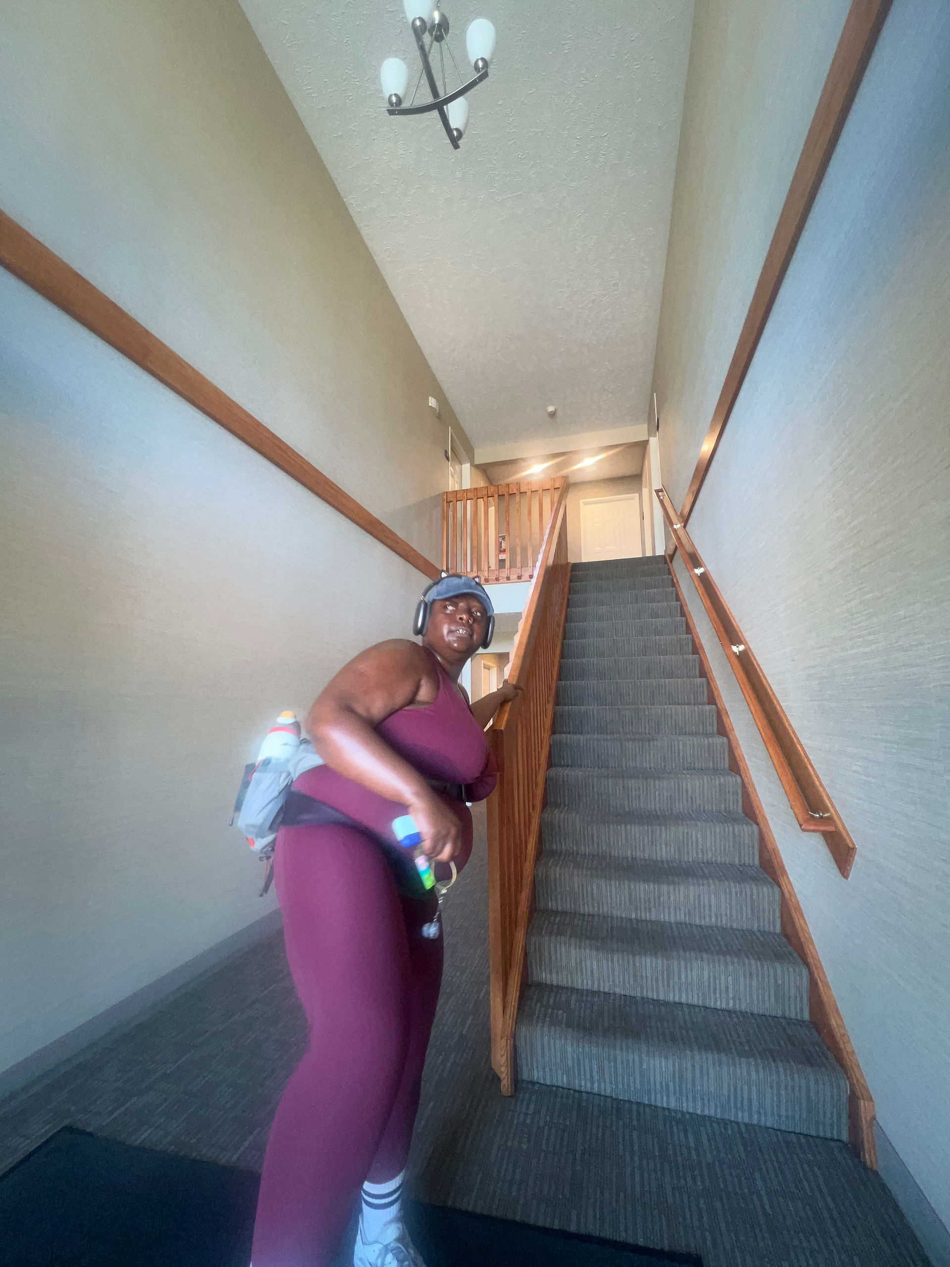 Kiing Curry reaches toward the stair railing at the end of a workout. Wearing the same burgundy workout set, headphones, hiking waist pack, and sacroiliac belt, they pause at the first step.