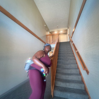 Kiing Curry reaches toward the stair railing at the end of a workout. Wearing the same burgundy workout set, headphones, hiking waist pack, and sacroiliac belt, they pause at the first step.