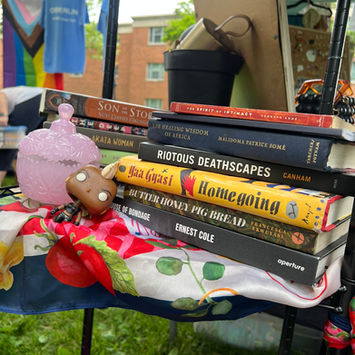 Stack of books and colorful objects arranged on a patterned table as part of a Bead + Cowrie/ The Lab traveling community center display.