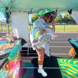 Person posing playfully inside a vendor tent with one foot raised toward the camera. Jewelry displays, books, holographic table coverings, and bright summer sunlight fill the booth.