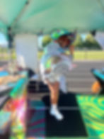 BespokeCurry at an outdoor pop-up wearing the white Spunge sneakers as part of their striped-and-blue Cosmos uniform, smiling while lifting one foot directly toward the camera so the sculptural outsole fills the foreground. Bead and adornment displays surround them inside the working field station.