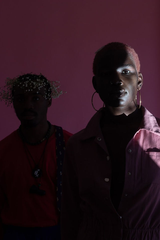 The same two Black models stand within deep magenta shadow. A sharp beam illuminates half of the coral-haired model’s face and mauve jumpsuit while the flower-crowned model recedes into darkness behind them.