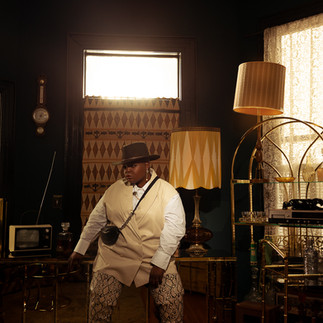 Black queer creative seated inside vintage-inspired room set in Adora house with warm lighting, patterned wallpaper, and retro furniture during narrative-driven fashion editorial production.