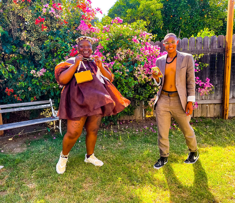 Kiing stands beside Siir in a sunlit garden overflowing with pink, red and white flowers. Kiing wears a voluminous brown off-the-shoulder mini dress, leopard-print cap, white sneakers and a small yellow purse, while the other person wears an open gray suit with black sneakers. Both smile toward the camera. Kiing’s fat body takes up substantially more physical space within the frame, making bodily scale and proportion visually legible without isolating either person.