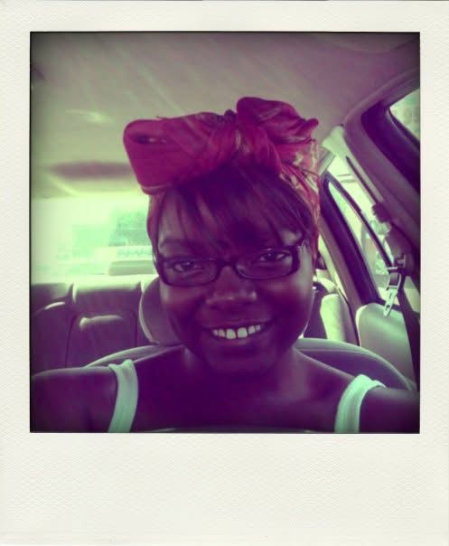 A purple-and-green-tinted Polaroid-style self-portrait of Kiing smiling inside a car. They wear rectangular glasses, a white tank top and a large red headwrap tied into a sculptural bow above their forehead.