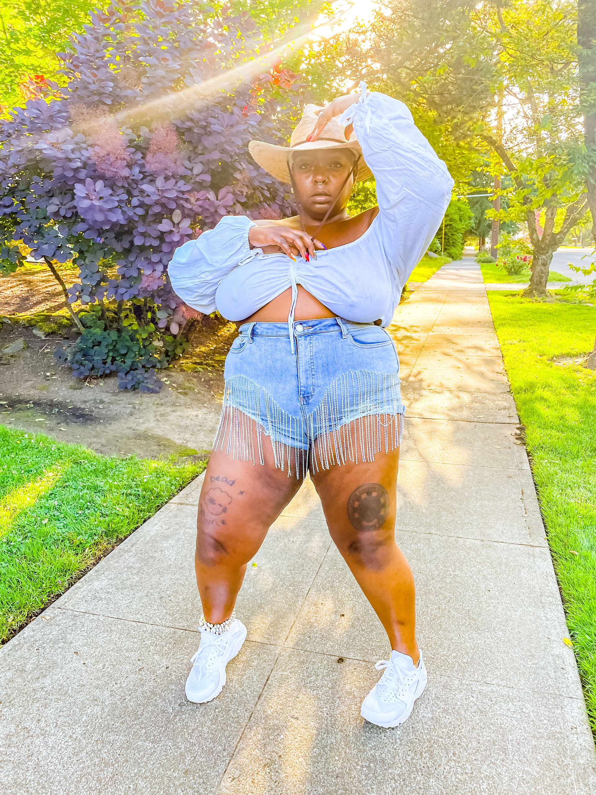  Kiing Curry stands on a sunlit sidewalk wearing a white gathered blouse, rhinestone fringe denim shorts, a straw cowboy hat, and white sneakers with sunlight streaming through nearby trees.