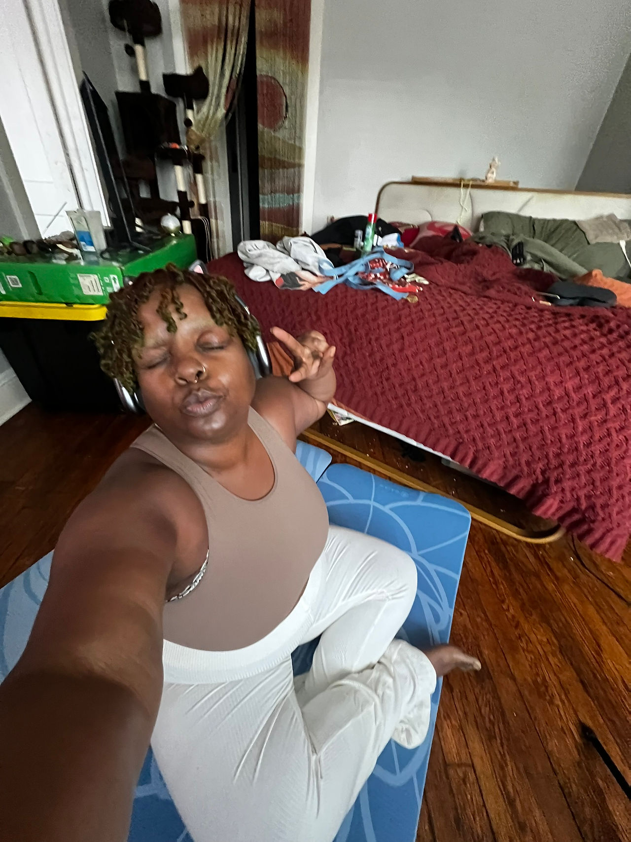 elf-portrait of Kiing Curry seated cross-legged on a blue yoga mat with eyes closed and over-ear headphones on. They wear cream ribbed pants and a taupe tank top while making a peace sign. A lived-in bedroom surrounds them with an unmade bed, cat tree, and everyday objects visible in the background, documenting a quiet moment of practice before surgery in 2025.