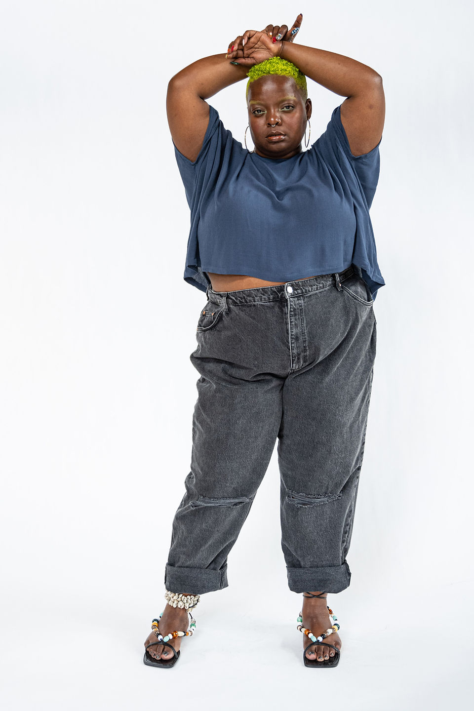 Kiing Curry poses in a blue cropped top and rolled charcoal jeans with both arms crossed overhead against a white studio backdrop.