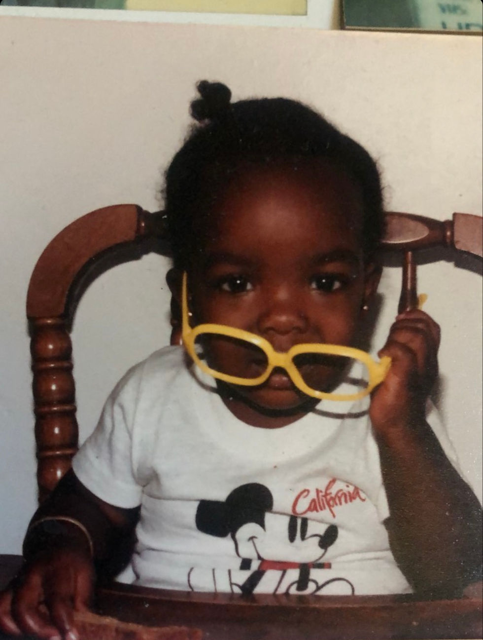 Childhood photograph of BespokeCurry as a young Black child seated in a wooden chair, holding bright yellow sunglasses to their face. Used to close “Mycelial Sankofa: Bridging the Network Again,” the image returns the essay from fungi, ecological relationship, Black embodied knowledge, and disrupted lineage to the child whose body was always perceiving, reaching, and communicating.