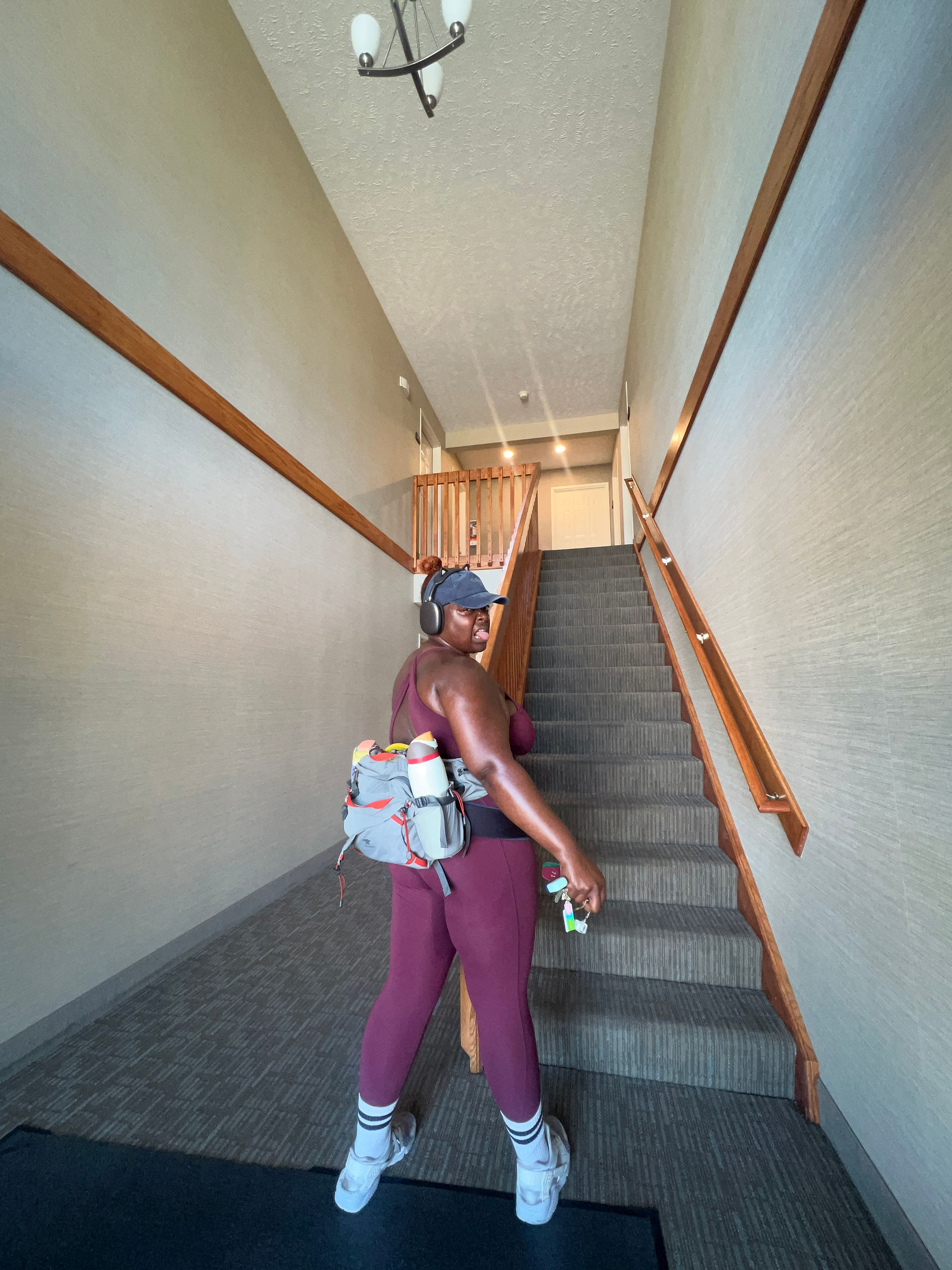 Kiing Curry stands at the base of a long apartment stairwell, looking back over one shoulder at the end of a workout. They wear a burgundy athletic set, white socks, sneakers, over-ear headphones, a baseball cap, a gray hiking waist pack with water bottles, and a sacroiliac support belt beneath the pack. The staircase rises dramatically behind them, emphasizing the challenge ahead.