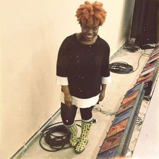 Black queer creative with bright orange textured hair smiling mischievously backstage during Cabaret rehearsal process while standing near stage equipment and lighting materials.