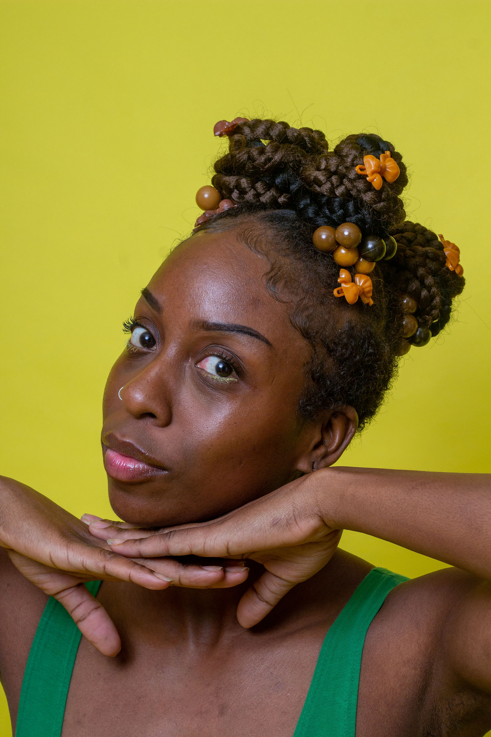 A Black model poses against a vivid yellow background with their chin resting theatrically across both hands. Their hair is arranged into large braided buns decorated with round wooden beads and small amber barettes and hair bobbles..