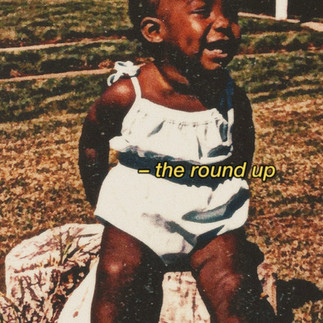 Photograph of a Black toddler sitting outdoors on a tree stump wearing a light-colored romper and white shoes.