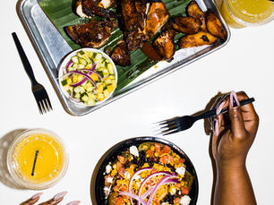 Overhead photograph of jerk-style glazed chicken and fried plantains on a banana-leaf-lined at Riddim n Spice in Nashville 2020. A metal tray with rice, cucumber salad and a yellow drink. Beside it, Kiing Curry’s hands frame a colorful grain bowl topped with corn, tomatoes, cheese and red onion.