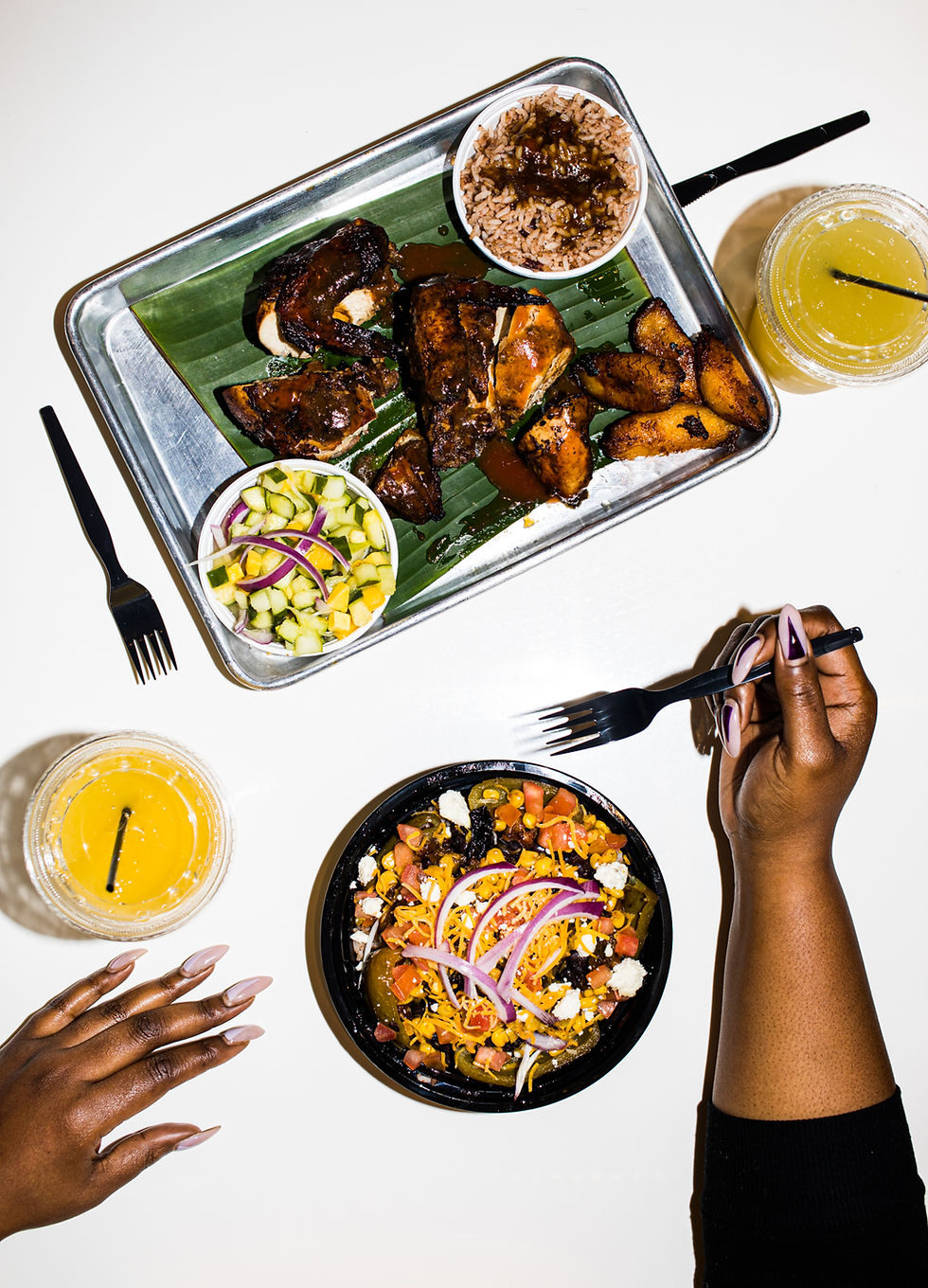 Overhead photograph of jerk-style glazed chicken and fried plantains on a banana-leaf-lined at Riddim n Spice in Nashville 2020. A metal tray with rice, cucumber salad and a yellow drink. Beside it, Kiing Curry’s hands frame a colorful grain bowl topped with corn, tomatoes, cheese and red onion.