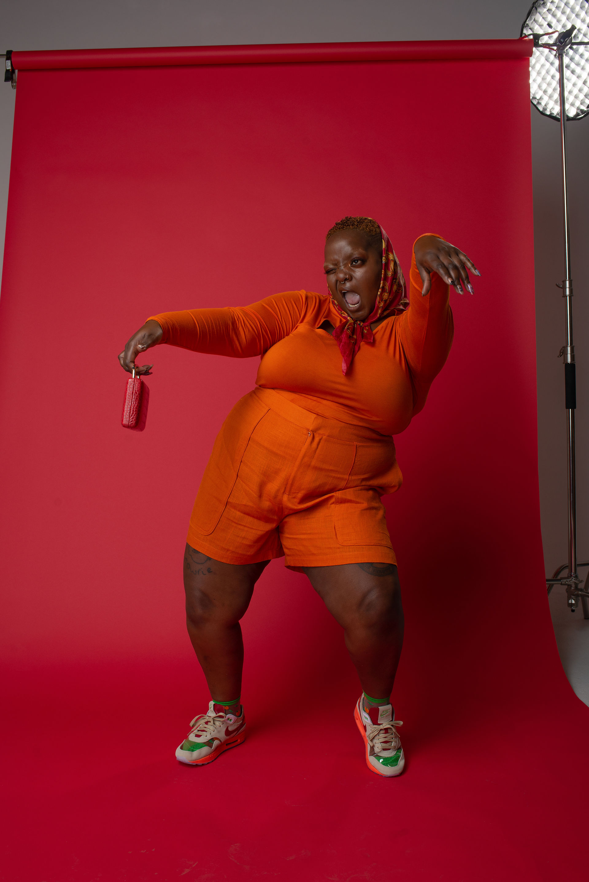 Black queer creative in oversized orange textured outfit posing dynamically while holding beverage can against vivid red backdrop during editorial fashion shoot.