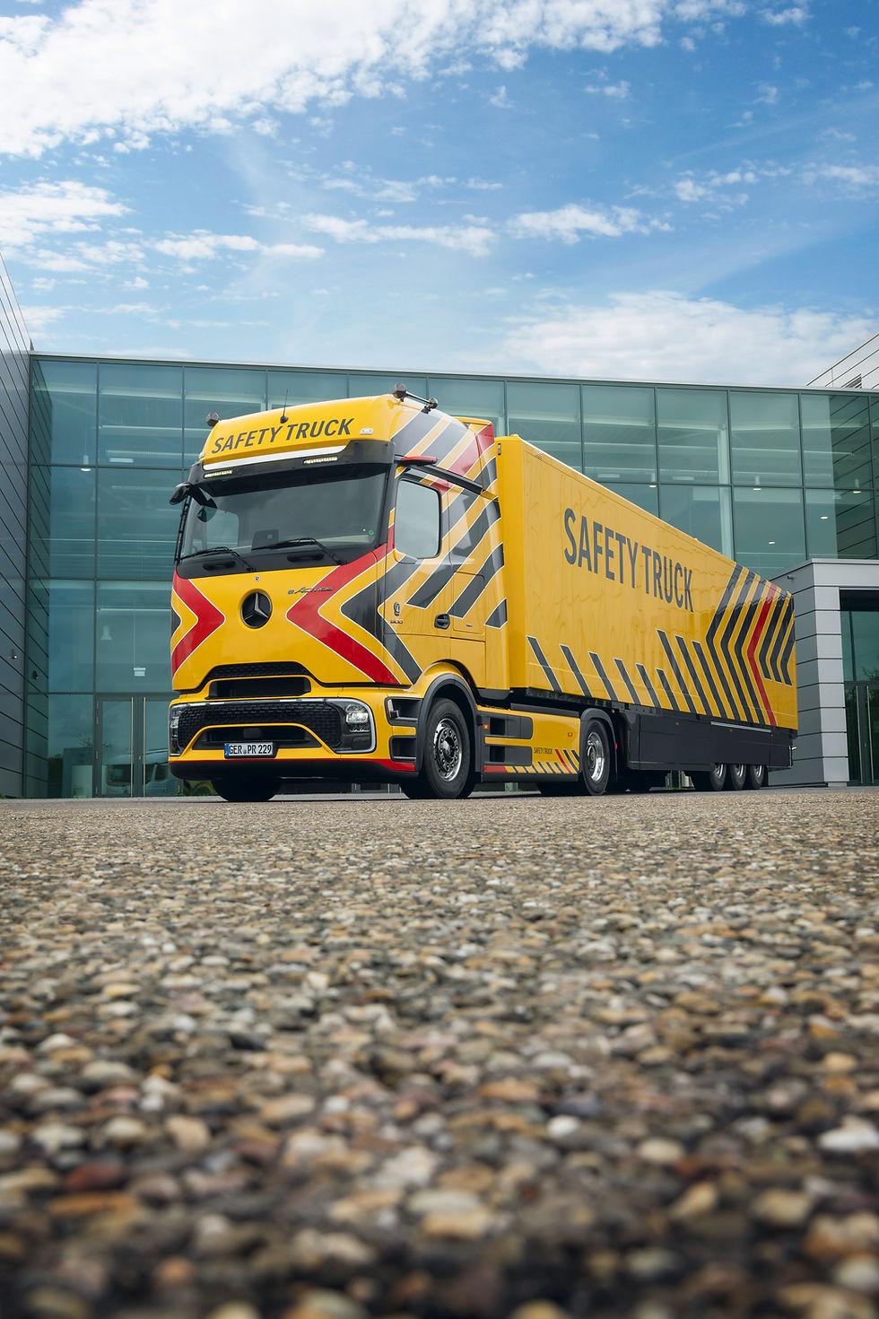Gelber Mercedes-Benz Lastwagen mit "SAFETY TRUCK" Schriftzug, vor modernem Gebäude, blauer Himmel. Markante rote Streifen, Betonboden.