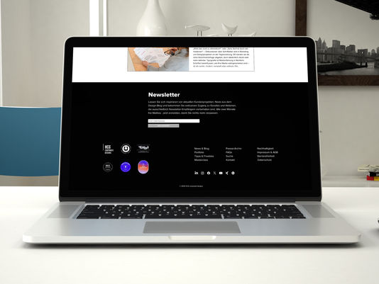 Laptop auf weißem Tisch mit offenem Newsletter-Formular im schwarzen Hintergrund. Blaue Stühle, Bücher und ein schwarz-weißes Bild im Hintergrund.