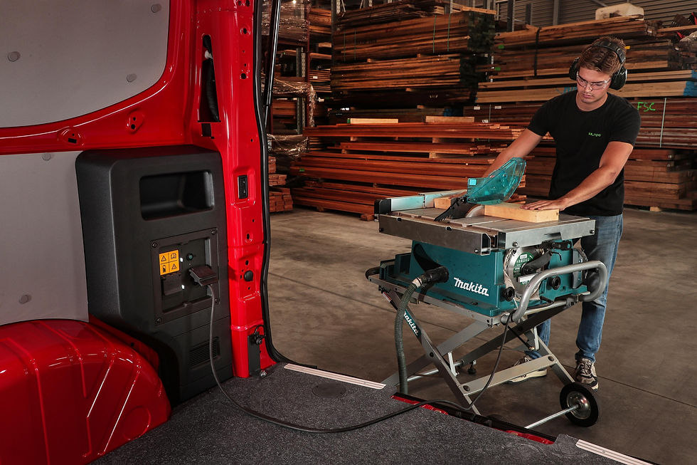 Mann mit Gehörschutz sägt in einer Holzwerkstatt Bretter an einer Makita-Tischkreissäge; im Vordergrund roter Transporter.