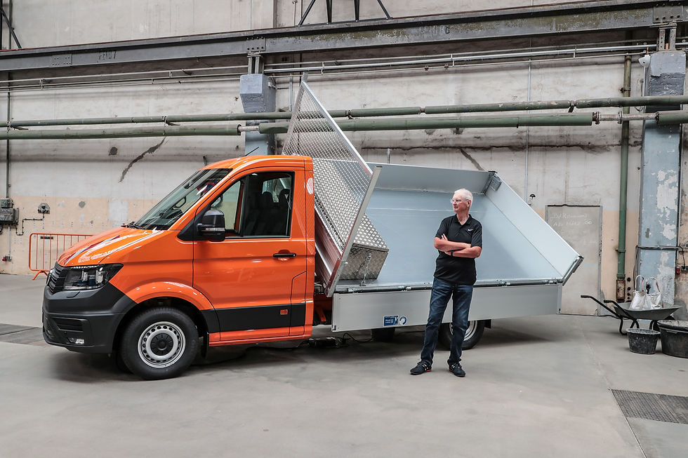 Orange Kastenwagen mit offener Ladefläche in einer Industriehalle; Mann steht daneben, Arme verschränkt.