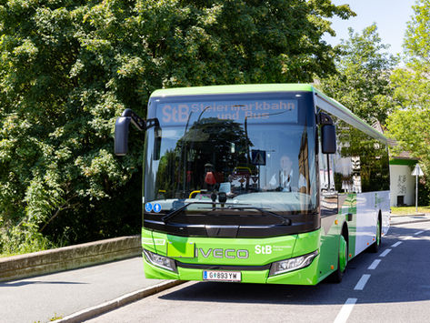 Grüner StB-IVECO-Linienbus auf sonniger Straße am Waldrand; digitale Anzeige mit SB-Text, ruhige Szenerie.