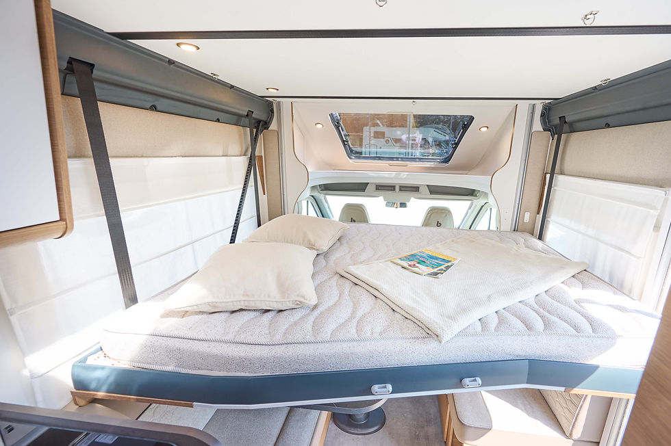 Helles Wohnmobil-Schlafzimmer mit Bett, Kissen und Magazin, Blick durch das Dachfenster ins Fahrerhaus.