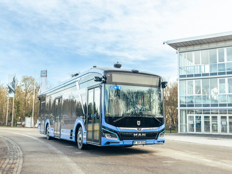Ein blauer autonom fahrender MAN Elektro-Bus fährt auf einer Straße vor einem modernen Gebäude. Der Himmel ist klar. Rückspiegel reflektieren Bäume. Seriennummer sichtbar.