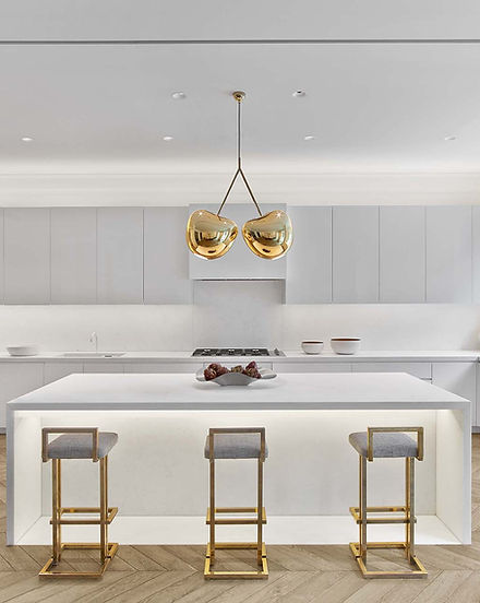 Modern, open kitchen with a large central island and seating. Brass cherry pendant lights hang over the island, which is also illumated with under-cabinet lighting. There are herringbone floors, and the entire space is white.