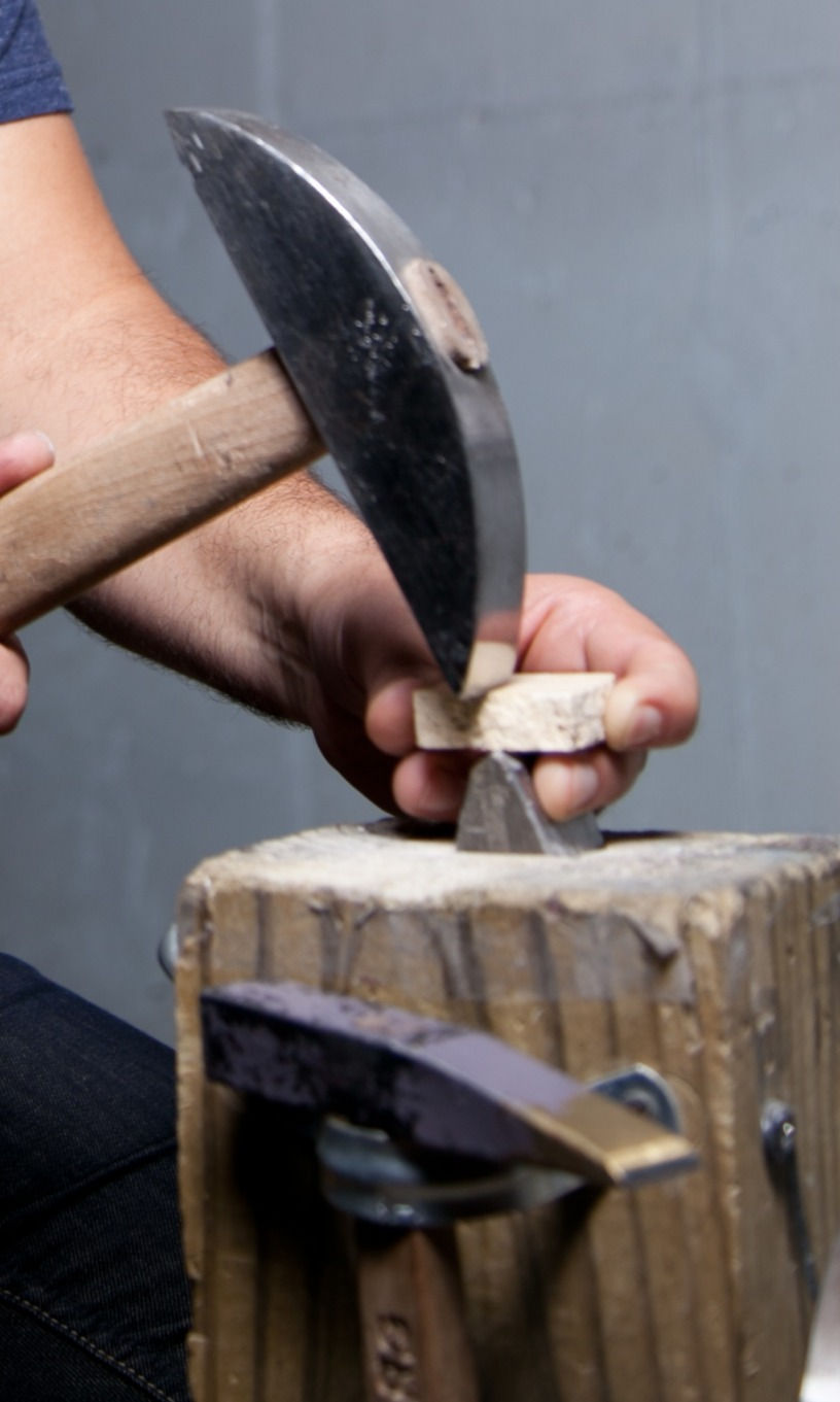 Close-up of hands using a mosaic hammer and hardie to cut a piece of light-colored stone into tesserae on a wooden work surface.
