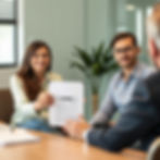 A smiling woman holds a "Loan Approved" document, sitting beside a man in an office setting with a plant in the background.