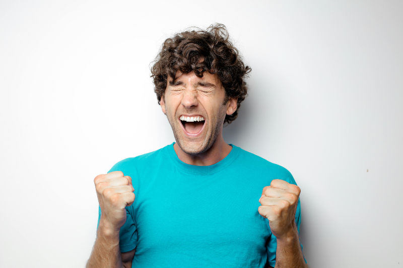 Man in a bright blue shirt expresses excitement with clenched fists, eyes closed, and mouth open against a plain white background.