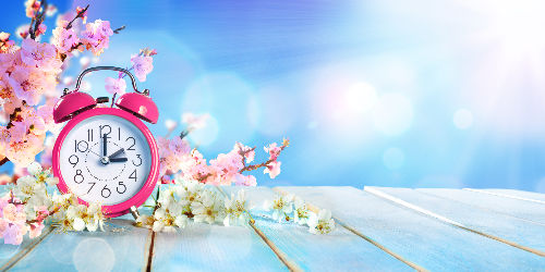 Pink alarm clock with cherry blossoms on a wooden table under a sunny blue sky. Bright, serene spring atmosphere.