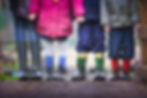 Four children standing side by side on a muddy path, each wearing colorful rain boots. They are holding hands, showcasing unity and friendship. Only their lower halves are visible.